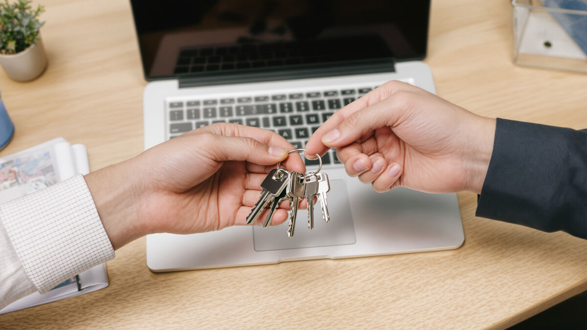 One hand passing a set of keys to another across a desk beside a laptop